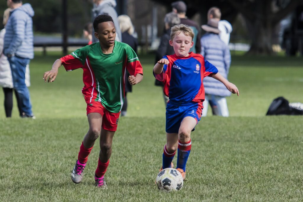 Two boys in school uniforms playing football outdoors, one about to kick the ball while the other runs to intercept, with a grassy field.