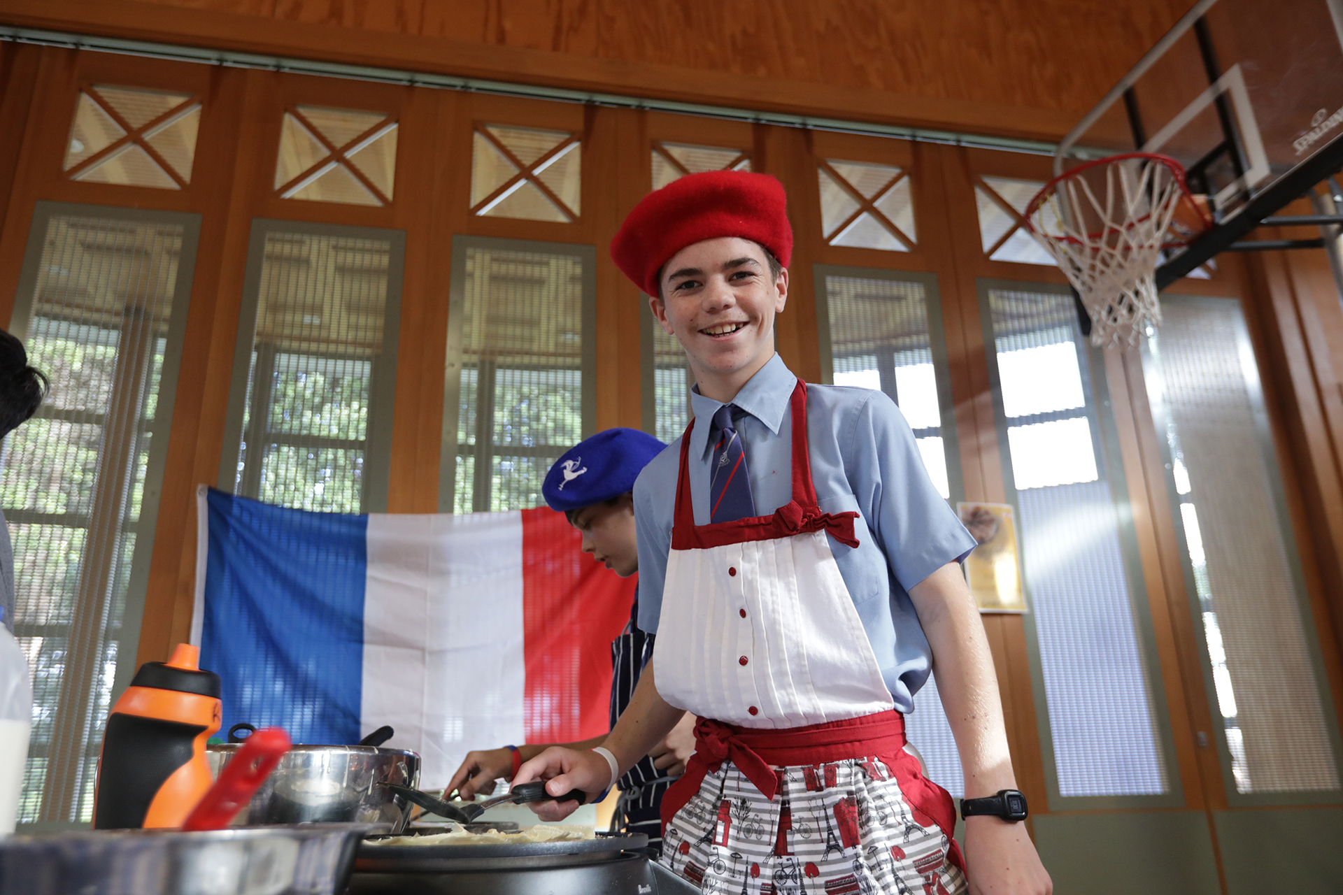 A boy wearing a French-style cap cooks a crepe, smiling and flipping a pan