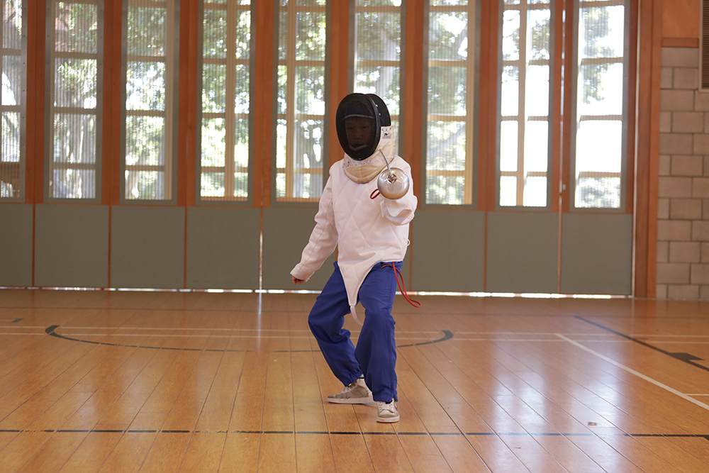 Student fencing indoors at Wellesley College Hall, wearing protective gear and holding foils