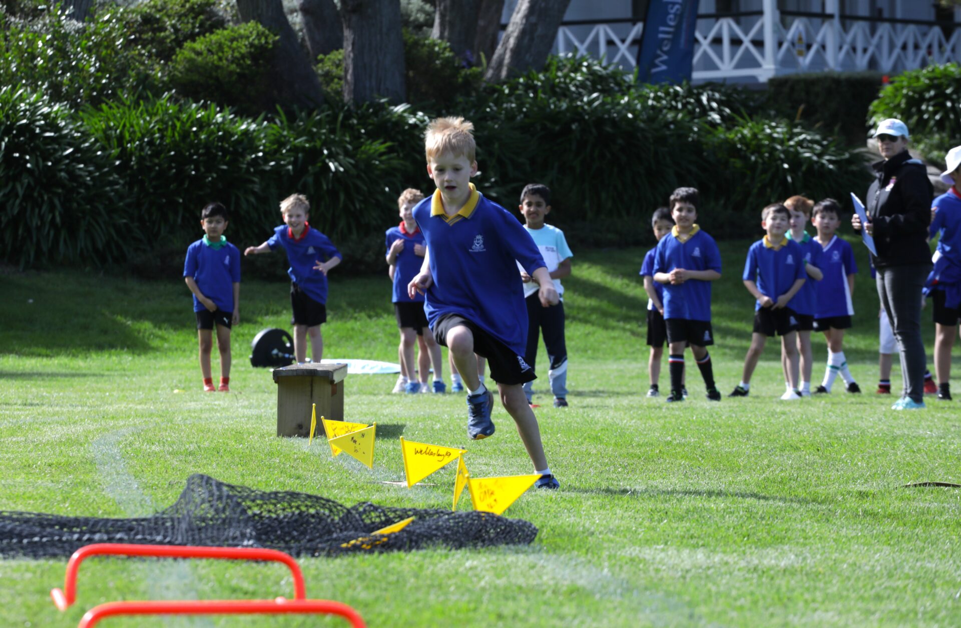 A Wellesley boy in sports uniform runs across the field while other boys in the background cheer him on