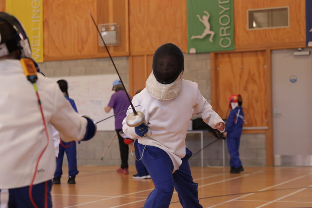 Two people wearing fencing gear and helmets face each other in a gymnasium, holding swords. Other participants and instructors are visible in the background near colorful banners and wooden walls