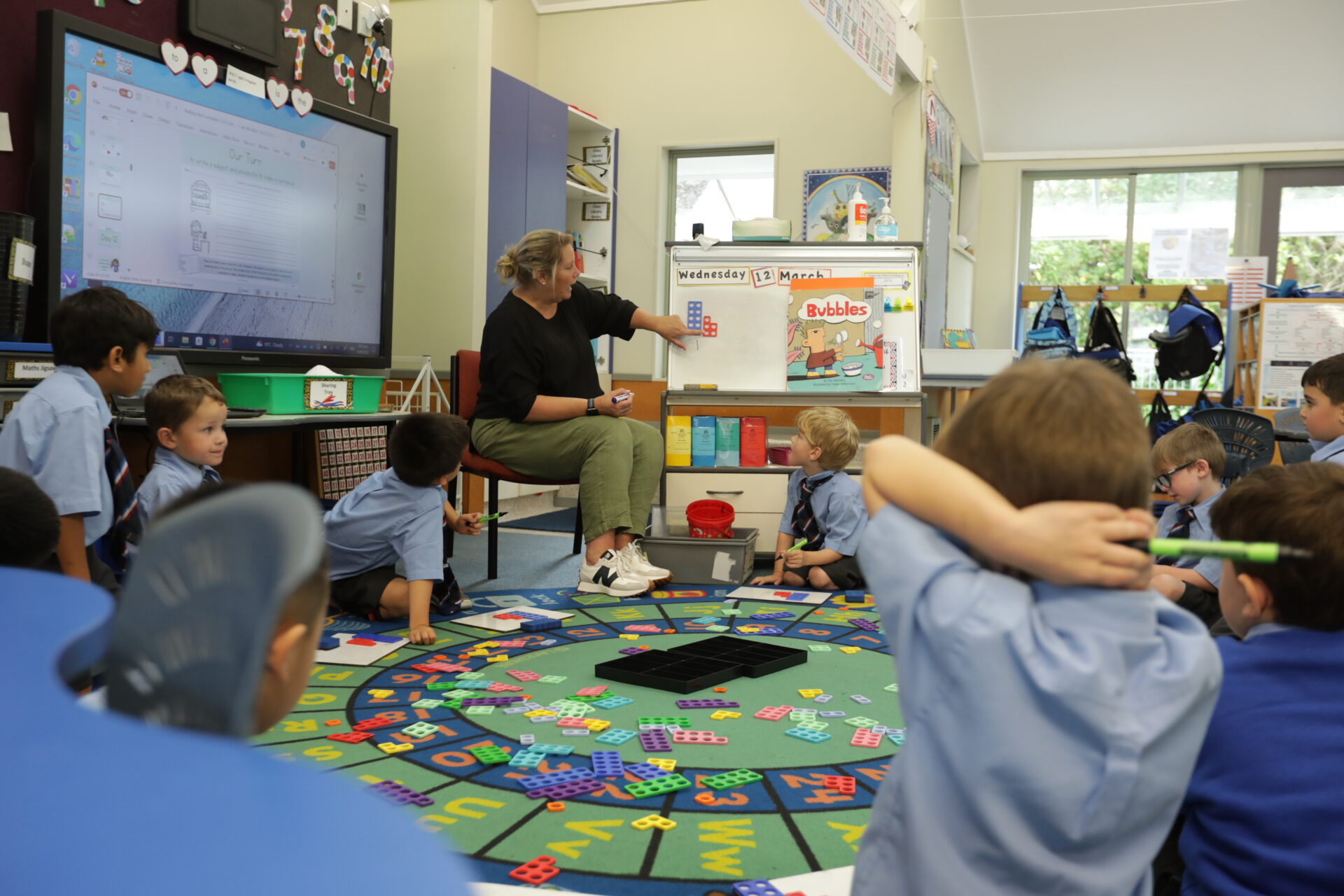 A teacher sits at the front of a colorful classroom, pointing at a chart while young students in blue uniforms sit on the floor, watching and listening attentively. Educational materials are spread out on the carpet