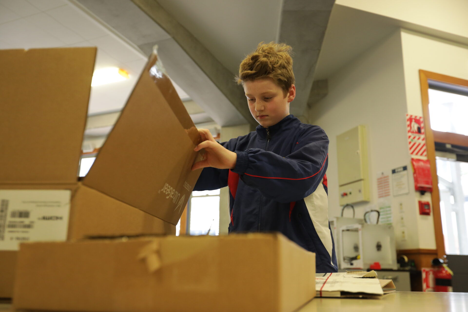 A young boy wearing a blue jacket opens a cardboard box on a table in a well-lit indoor setting, with other boxes and office supplies visible around him
