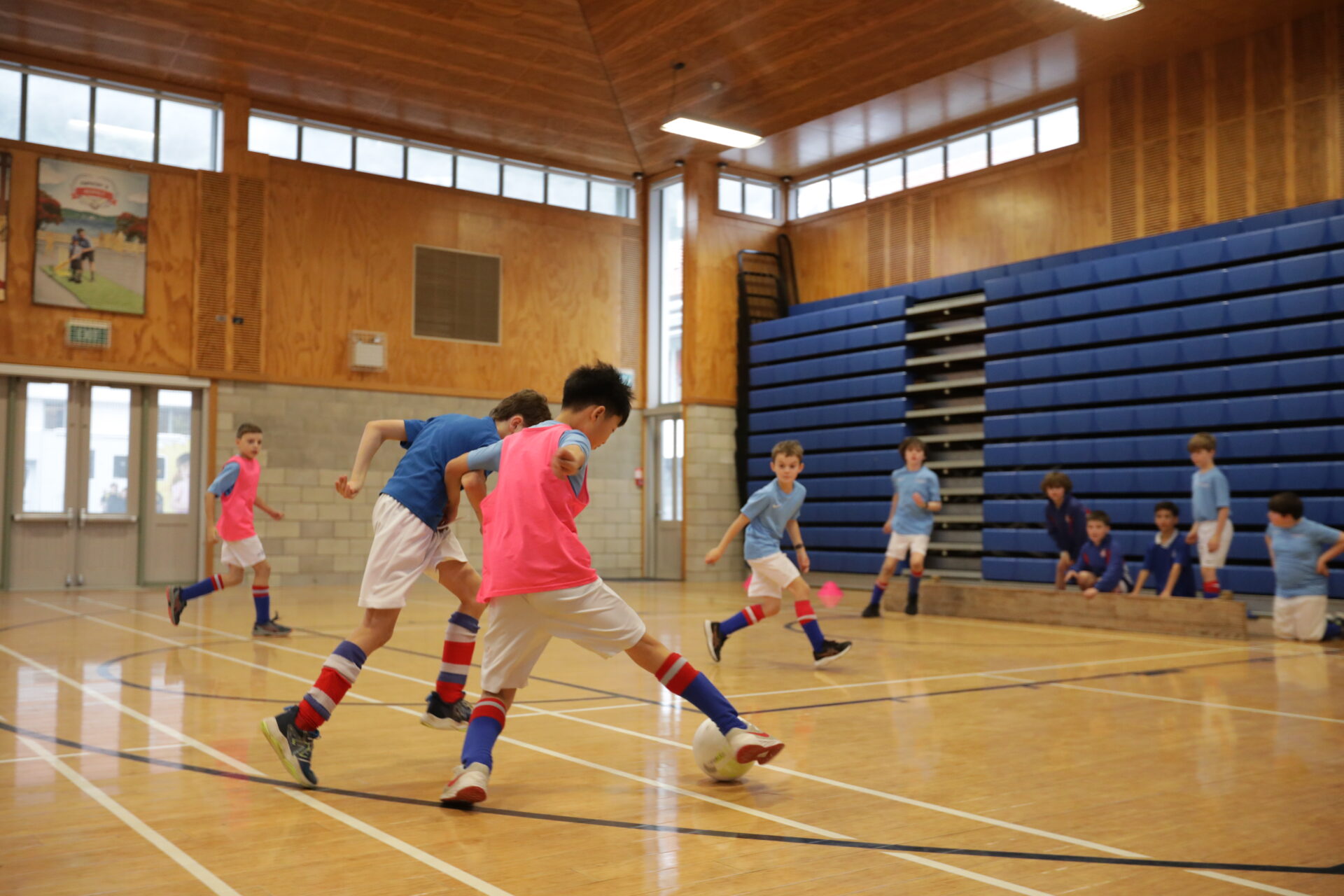 Boys play an indoor football game in a gym. Two teams, one in blue shirts and one in pink bibs, compete for the ball while others watch from the bleachers