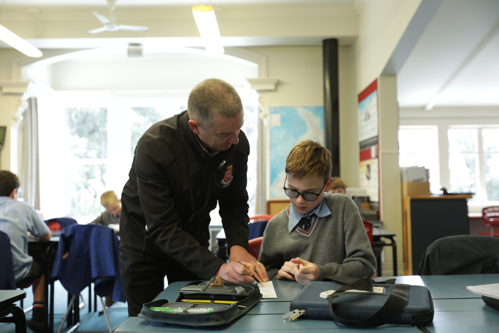 A teacher leans over to help a student with glasses, who is writing at a desk in a classroom. Other students are visible working in the background. School bags and supplies are on the desk