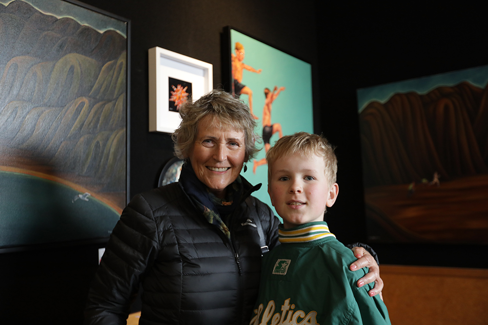 A boy stands with his grandmother, smiling together in front of Dean Proudfoot’s artwork displayed in the background