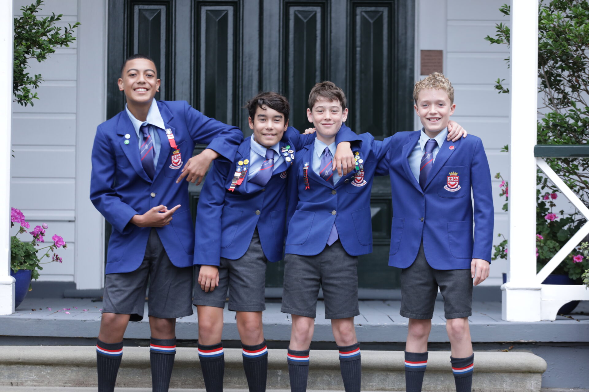 Four boys wearing blue Wellelsey College blazers, ties, and shorts stand smiling together on a porch, arms around each other in front of a green door and white building