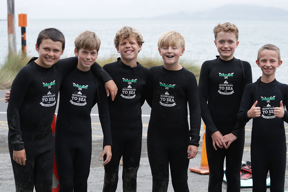 A group of boys at Days Bay prepare to go diving, standing together on the shore in their swim gear and looking excited