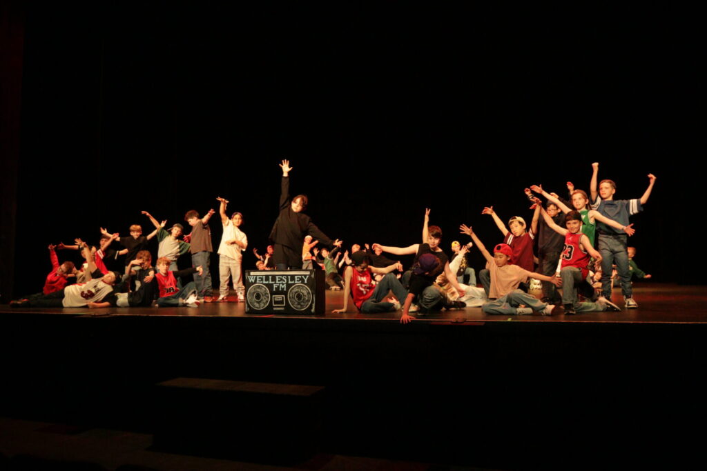 A group of children poses energetically on stage during a performance, with arms raised and expressive gestures. A sign reading Wellesley FM and a boombox are placed at the front of the stage