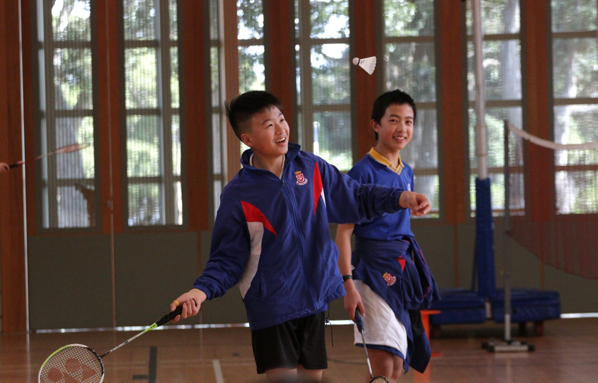 Two boys in sports uniforms play badminton indoors, smiling and looking up at a shuttlecock in midair.