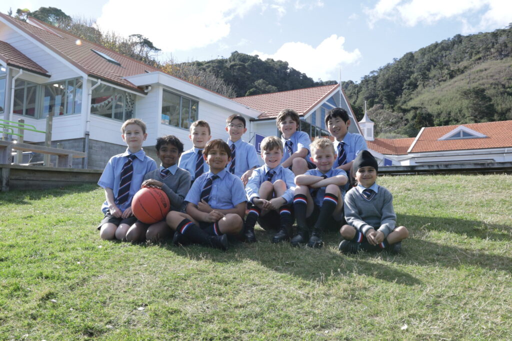 Boys in school uniform sitting on a grassy slope in front of a white school building, smiling, one boy in front holds an orange basketball